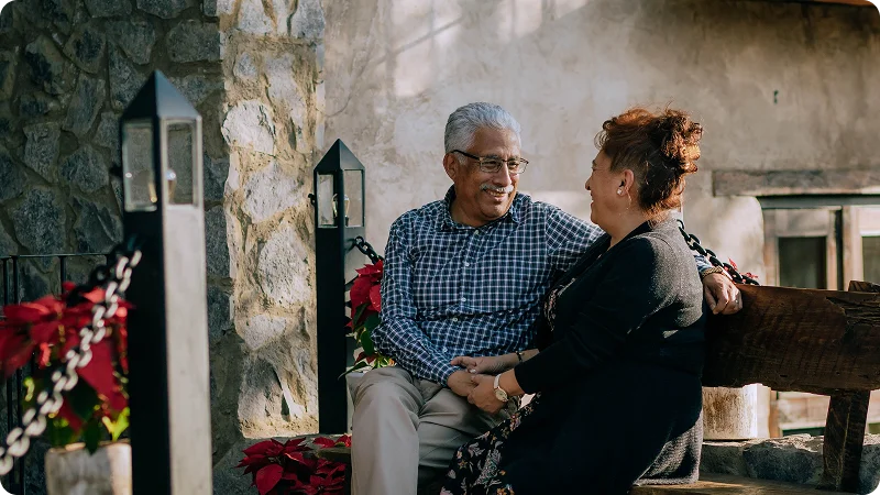 An older man and woman sitting on a wooden bench outdoors, smiling and looking at each other.