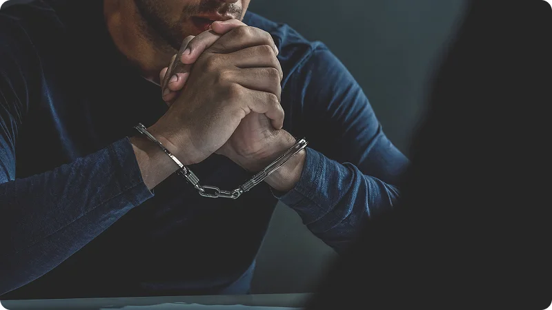 A man in a dark long-sleeved shirt sitting with his handcuffed hands clasped together in front of him.