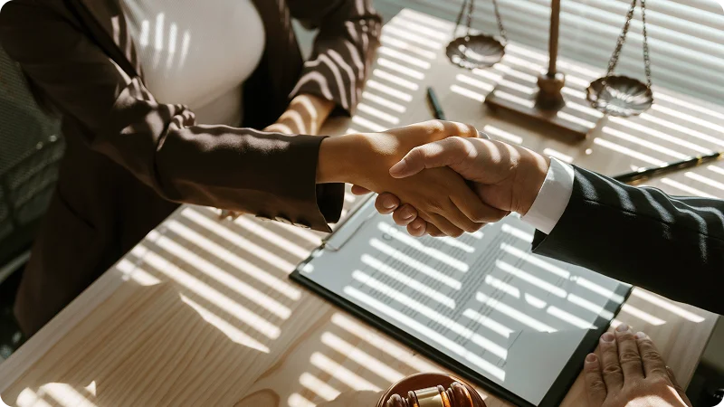 Two professionals shaking hands over a desk with legal scales in the background.