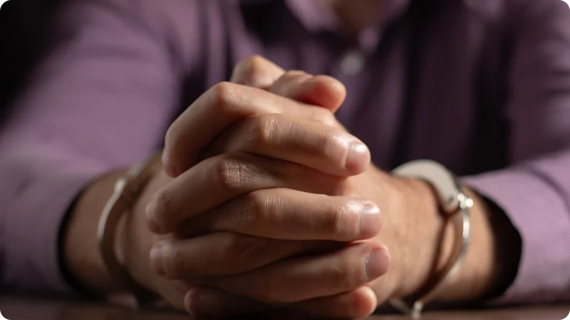 A person wearing a purple shirt with their hands clasped together while wearing handcuffs.