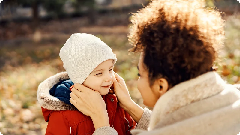 A woman with curly hair smiles while looking at a young child in a red jacket and white beanie in an autumn.