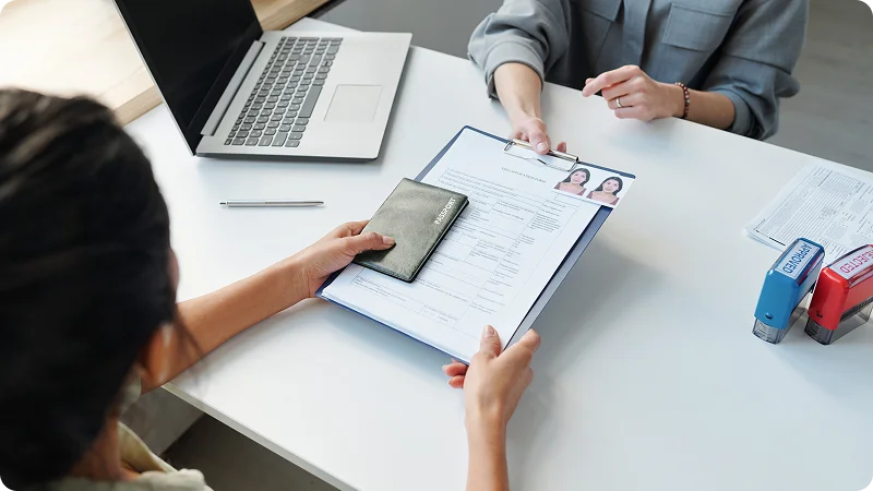 A person handing a passport and a document with American flag icons to an official at a white desk with a laptop and stamps.