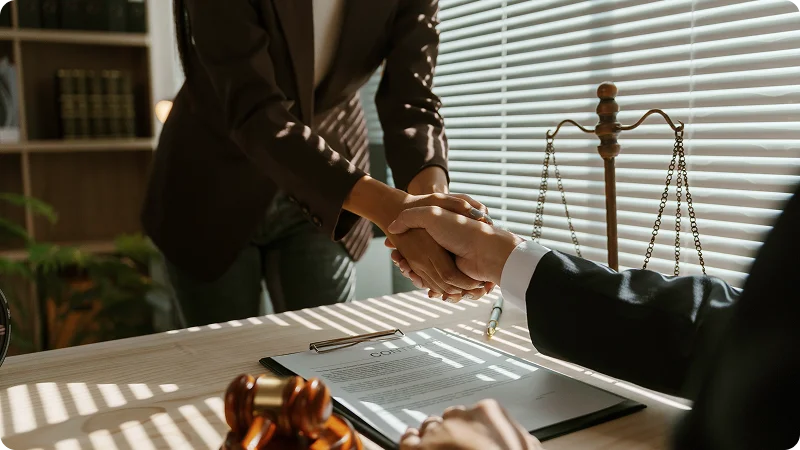 People shaking hands over a desk with a gavel and scales of justice.