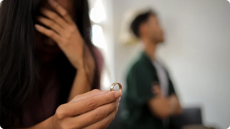 Distressed woman holding a wedding ring with a blurred man in the background.