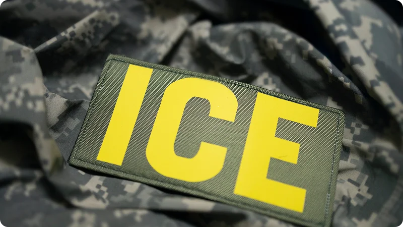 A green tactical patch with the word "ICE" in bold yellow letters, resting on camouflage fabric.