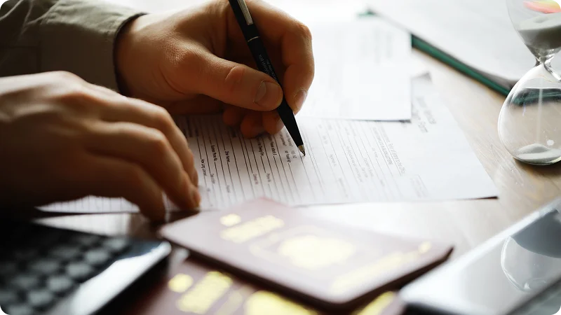 Hands writing on an official document with a passport and hourglass nearby.