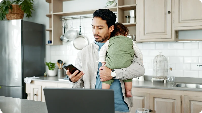 A man standing in a kitchen holding a sleeping baby while looking at his smartphone.