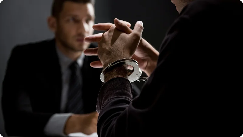 A man in a suit sitting across from another person whose hands are cuffed and clasped on a table.