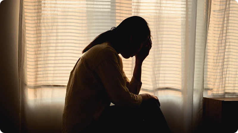 The silhouette of a woman sitting with her head in her hands in front of a window.