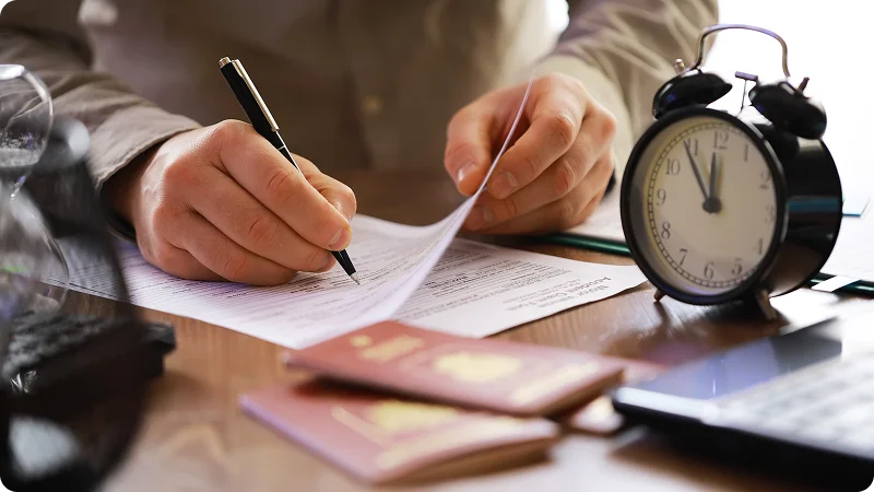 Hands filling out paperwork with a passport and alarm clock nearby.