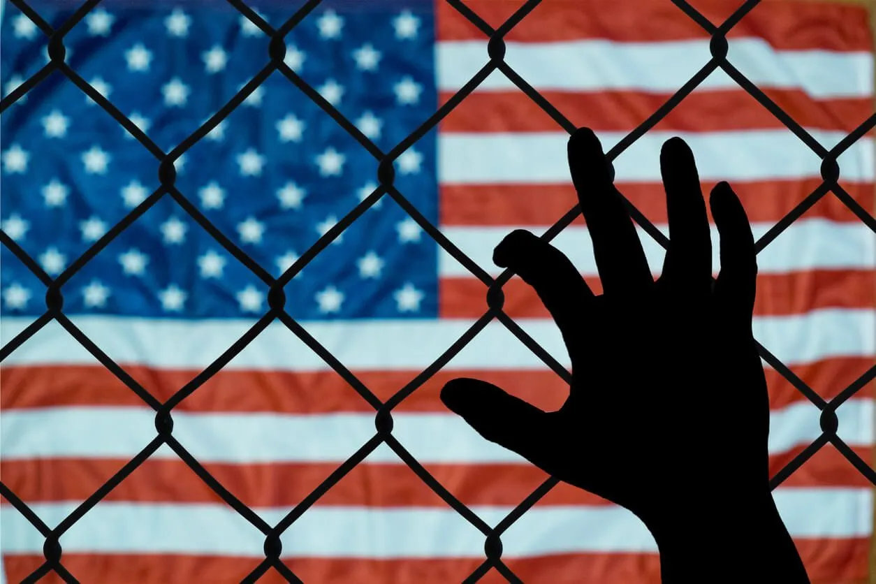 A silhouette of a hand reaching toward a chain-link fence with the American flag blurred in the background.