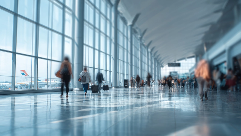 Blurry image of travelers with luggage walking through a modern airport terminal with large windows.