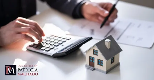 A person uses a calculator and a pen to review paperwork next to a small model house.