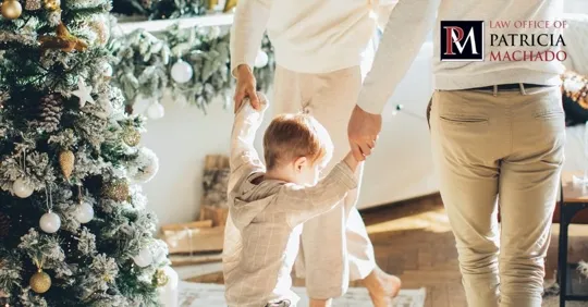 A young child holds hands with two adults while walking indoors near a decorated Christmas tree.