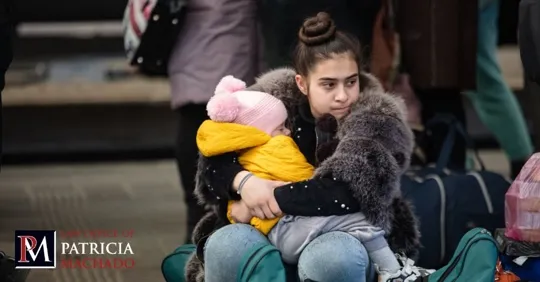 A young woman in a fur coat sits outdoors among luggage, holding a small child in a yellow jacket and pink hat.