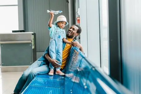 A man sits on a blue airport bench and smiles at a young child standing next to him holding a toy airplane.