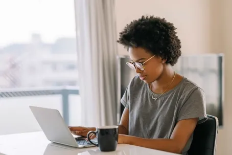 A woman with curly hair and glasses sitting at a table, focused on her laptop with a black mug nearby.
