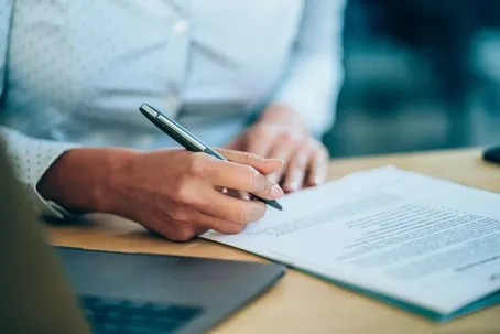 A close-up of a person's hand holding a pen while signing a formal document on a wooden desk.