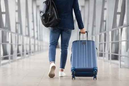 A traveler wearing a backpack and pulling a blue suitcase through a bright, modern walkway.