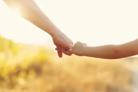 An adult and a child holding hands against a soft, golden-hour.