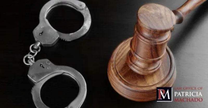 A set of silver handcuffs rests next to a wooden judge's gavel on a dark, reflective surface, representing criminal justice and legal proceedings. In the bottom right corner, there is a small logo for a law firm.